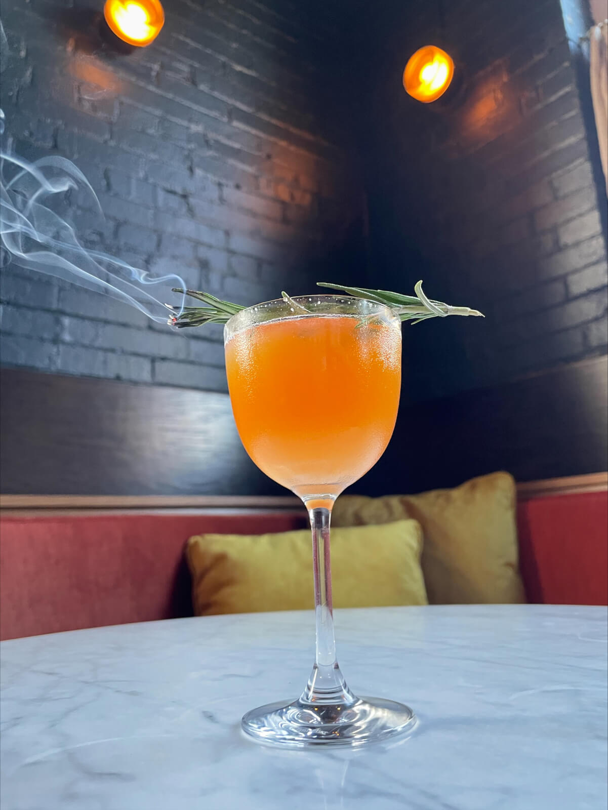 A cocktail in a glass garnished with a burning herb, placed on a marble table against a dimly lit background with red seating and yellow cushions.