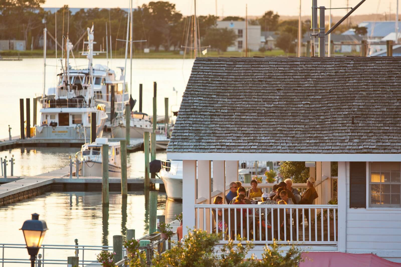A group of people enjoys a meal on the porch of a waterside building on the Crystal Coast, with several boats docked in the background at sunset. 