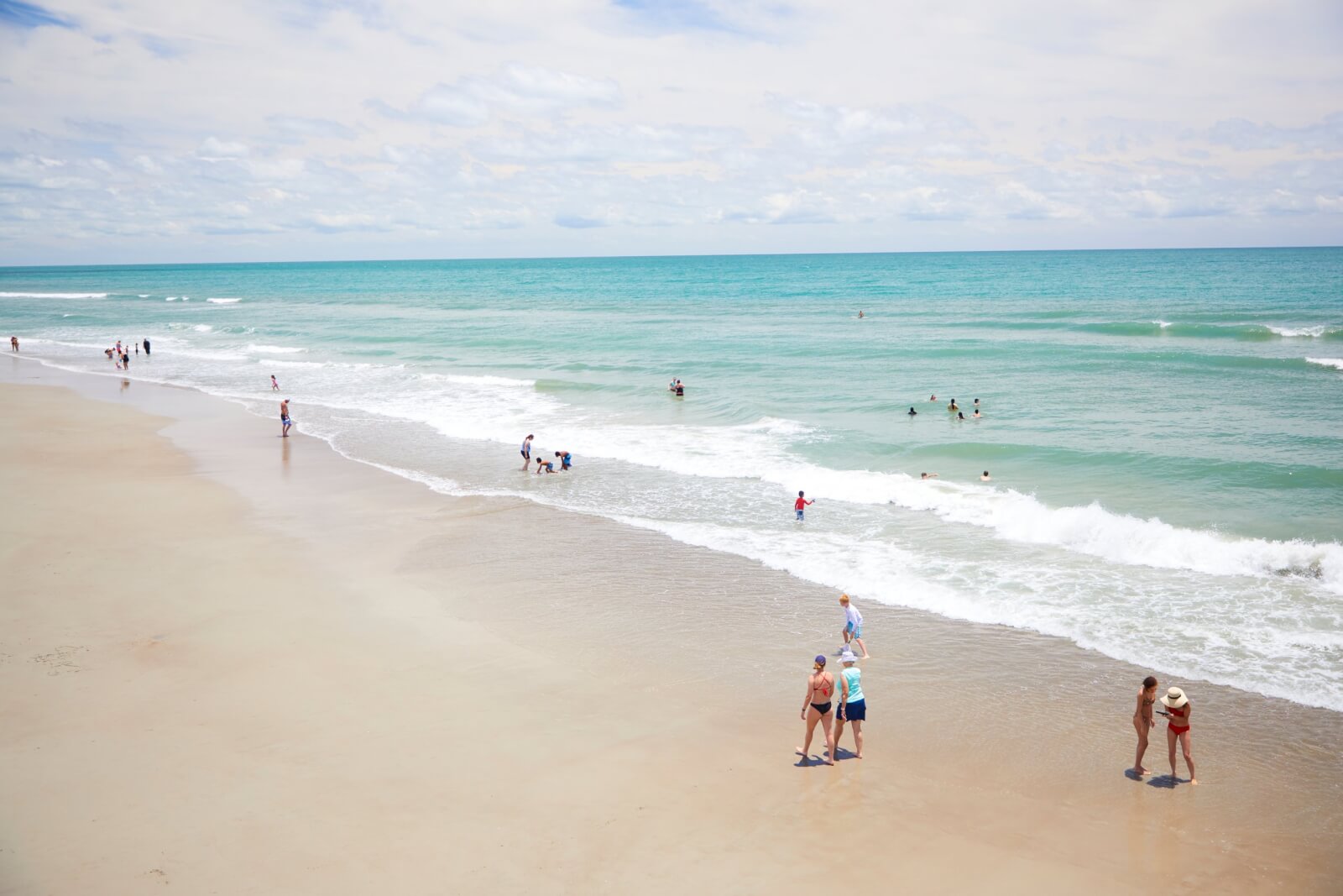 People are walking and playing along the shore of a sandy beach with calm waves under a partly cloudy sky.