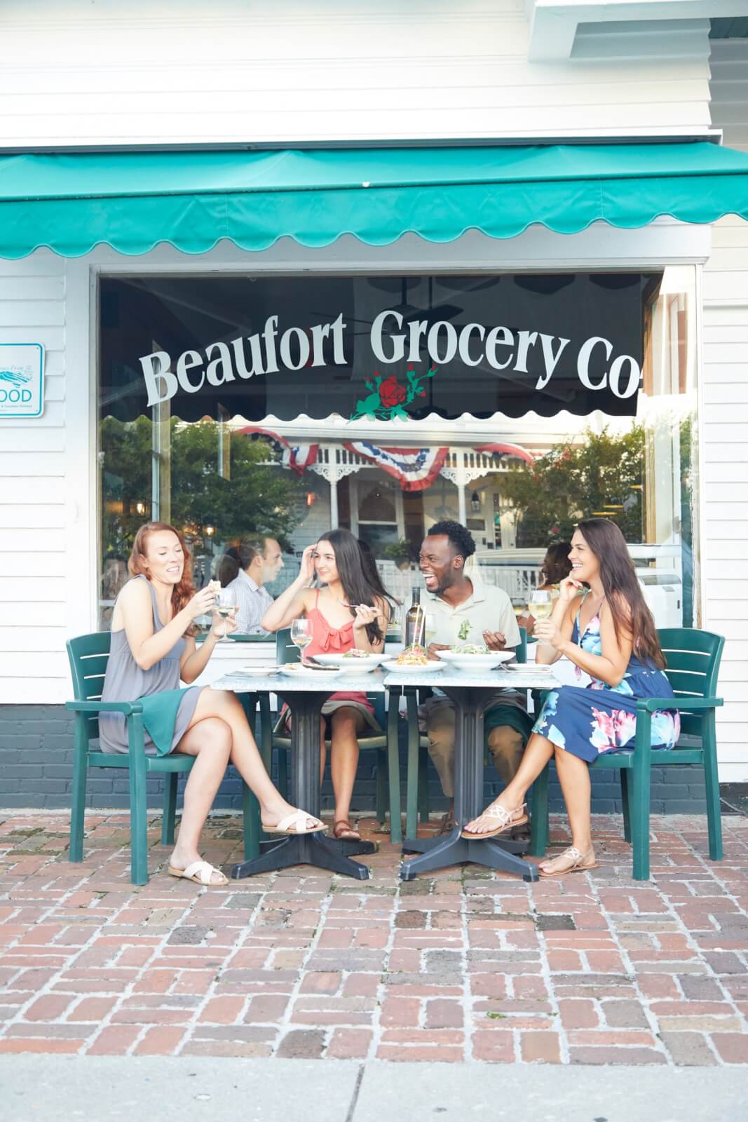 Four people sit at an outdoor table in front of Beaufort Grocery Co. on the Crystal Coast, enjoying drinks and conversing during their visit to North Carolina.
