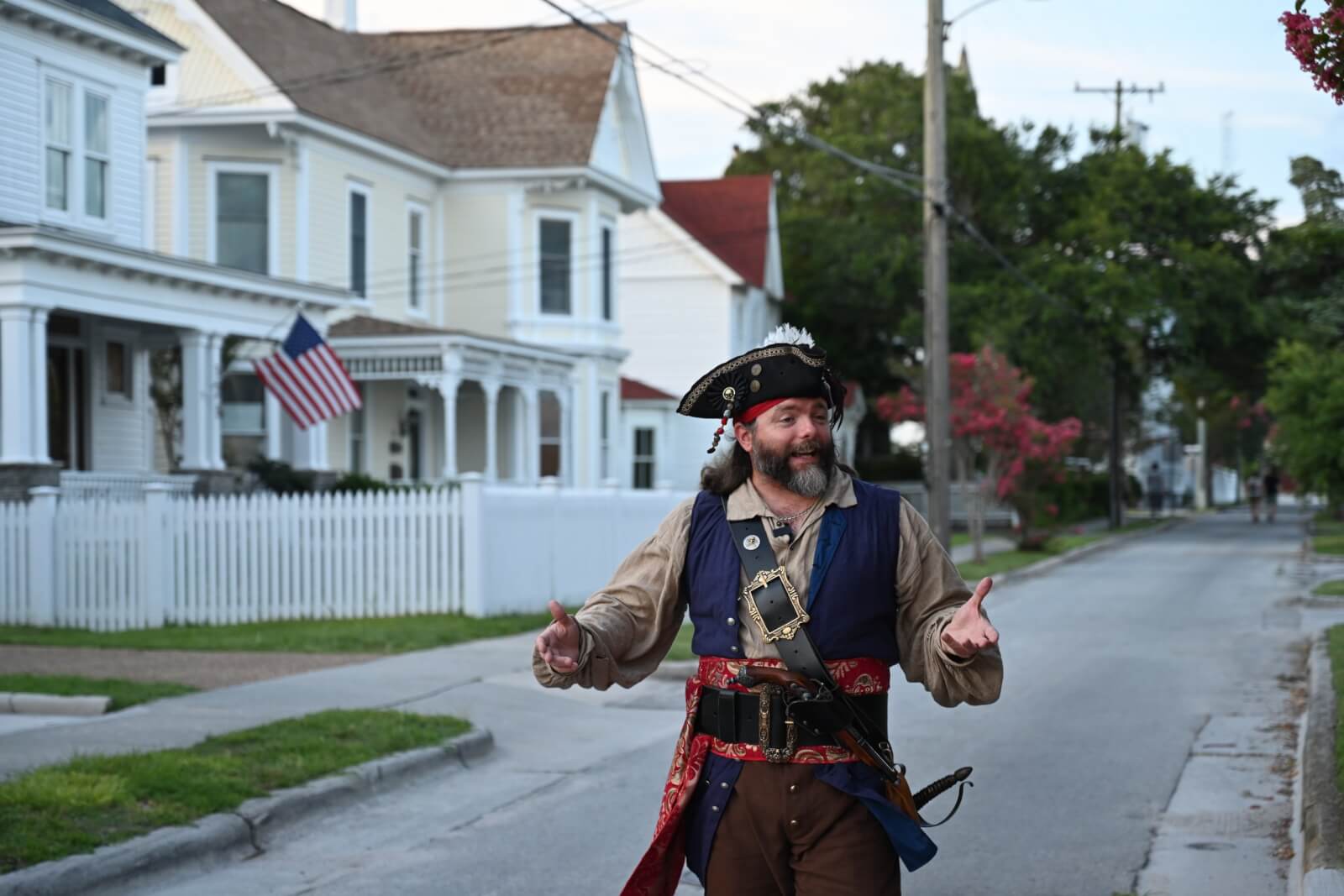 A man dressed as a pirate stands on a residential street with houses in the background, one of which has an American flag proudly displayed on its porch, capturing the spirit of North Carolina's Crystal Coast.