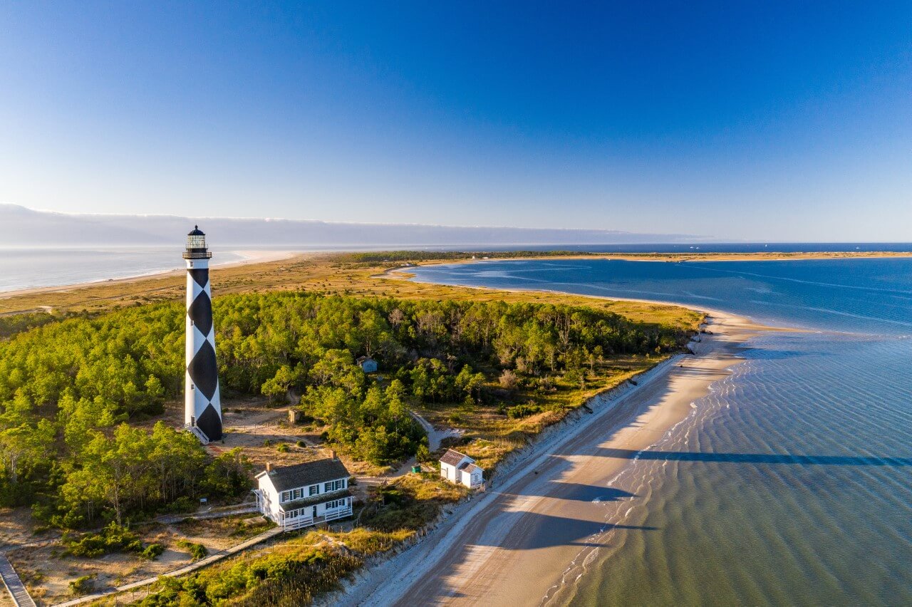 Aerial view of a lighthouse with a distinct black and white diamond pattern next to a white building on the sandy shoreline of North Carolina's Crystal Coast, surrounded by greenery with the ocean visible in the background.