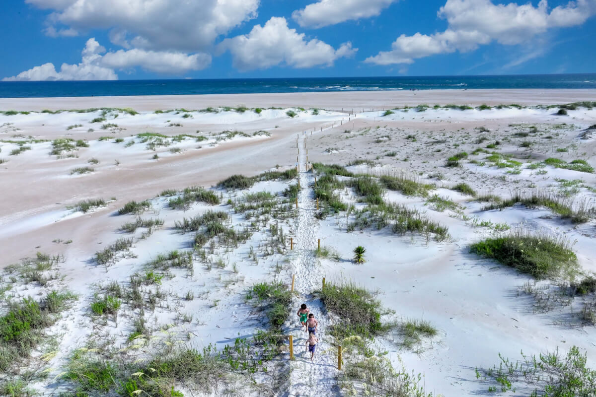Two people walk along a sandy path towards the beach under a blue sky with scattered clouds. The area, part of North Carolina's picturesque Crystal Coast, is surrounded by low vegetation and sand dunes. The shimmering ocean is visible in the distance, inviting them to visit its serene shores.