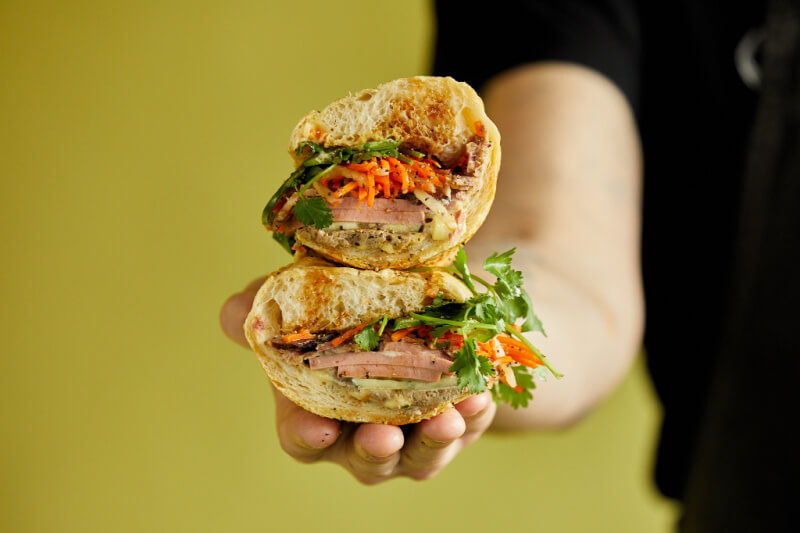 A hand holding a sandwich cut in half, featuring layers of meat, carrots, and fresh cilantro against a plain yellow background. 