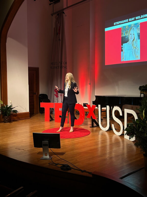 A person stands on a stage giving a TEDx talk. The background features a screen with the name Stephanie May Wilson and her photo, while large white TEDxUSD letters are displayed on the stage beside the speaker.