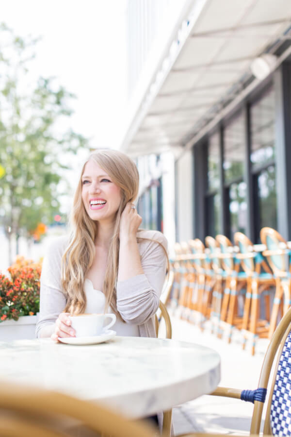 Stephanie May Wilson, a woman with long blonde hair, sits at an outdoor café table, smiling while holding a white cup and saucer. The background reveals empty chairs and tables, capturing the serene atmosphere that perfectly fits her joyful presence.