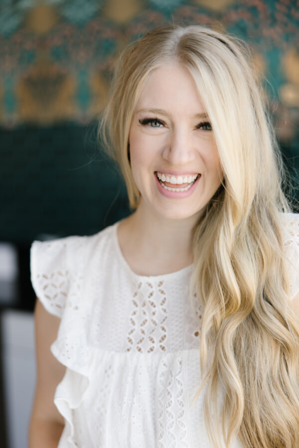 A smiling woman with long blonde hair, wearing a white, short-sleeved eyelet dress stands in front of a dark and colorful patterned background.