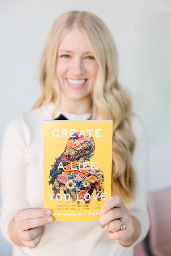A woman with long blonde hair is holding a yellow book titled "Create a Life You Love" by Stephanie May Wilson. She is smiling and wearing a white top.