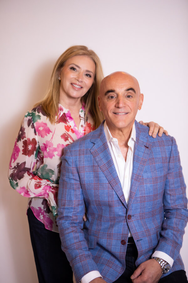 Theresa Scotto, a woman with long hair, wearing a floral blouse, stands next to a seated bald man in a blue plaid blazer. She rests her hand on his shoulder against a plain background.