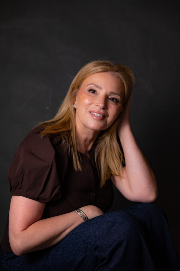 A woman with blonde hair, wearing a brown top and blue jeans, poses with a hand on her head against a dark background.