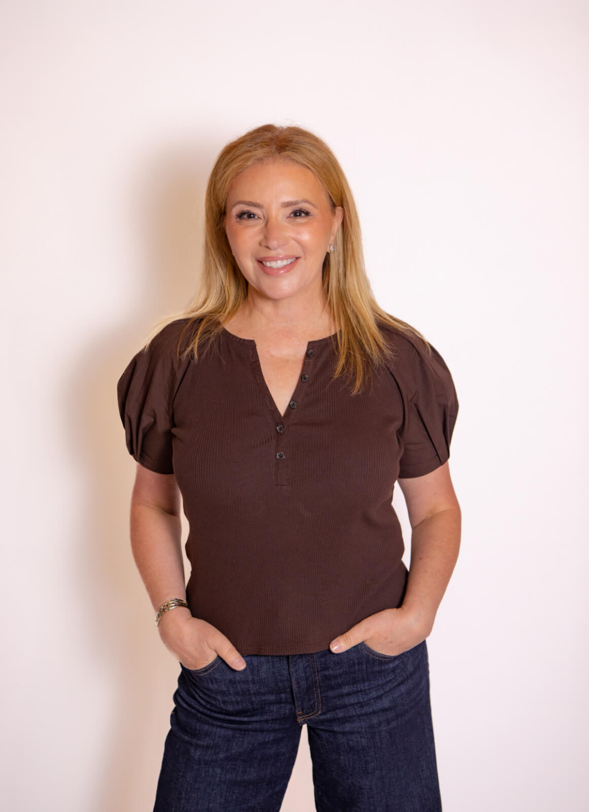 Theresa Scotto, a woman with long blonde hair, stands against a white background, wearing a brown top and blue jeans, with her hands in her pockets, smiling at the camera.