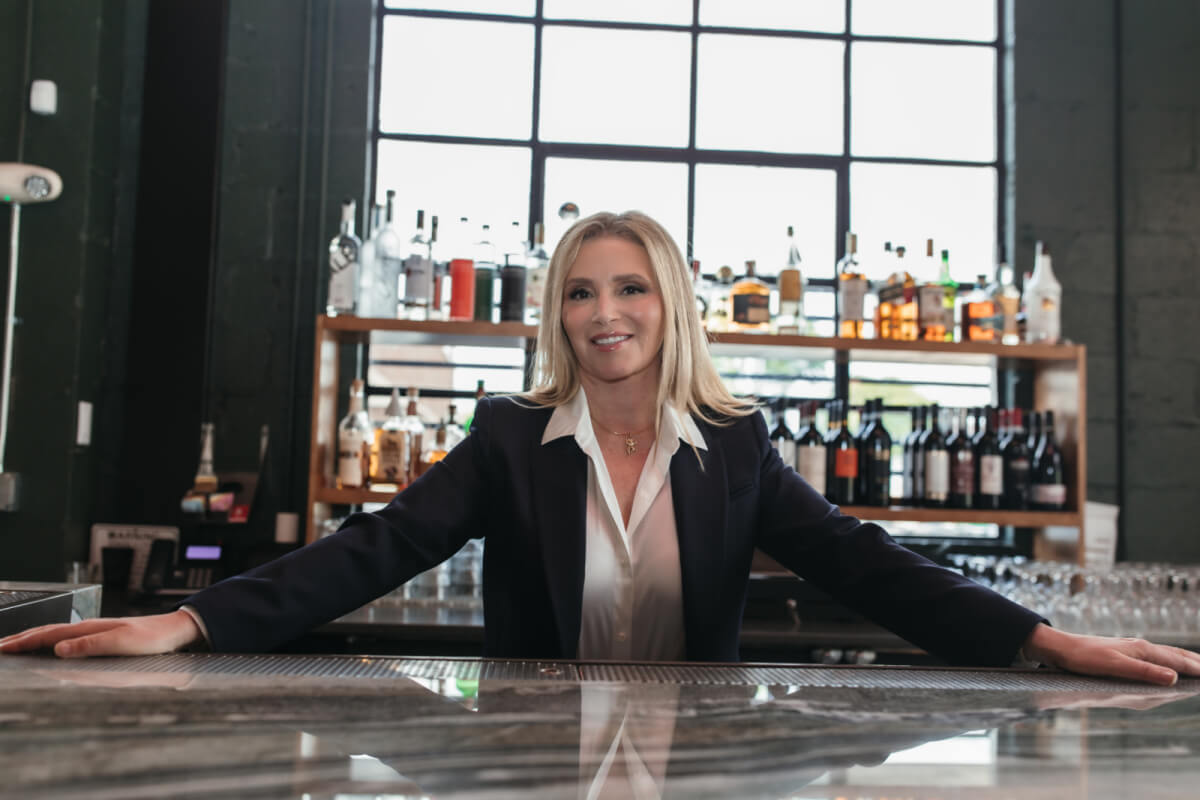 Theresa Scotto, a woman in a suit, stands behind a bar with bottles of alcohol on shelves in the background.