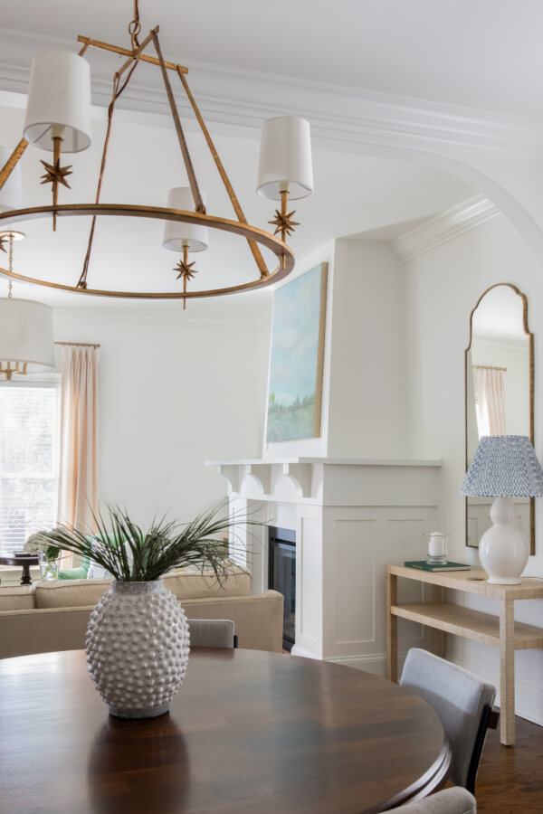 A Nashville Home living room designed by Elizabeth Burch Interiors features a round wooden table, a white vase with greenery, a large chandelier, a fireplace with a painting above the mantel, and a console table adorned with a lamp and mirror.