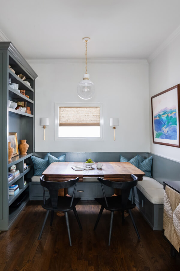 A cozy dining nook designed by Elizabeth Burch Interiors features a wooden table, black chairs, blue cushioned bench seating, built-in shelving, a pendant light, and a framed painting on a white wall—perfect for your Nashville Home.