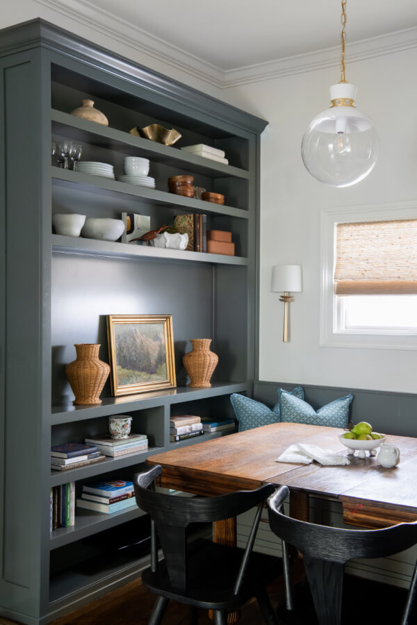 A cozy dining area with a wooden table and black chairs. A built-in shelf holds various decor items and books, creating a charming Nashville-inspired interior. A small framed painting and vases sit on a lower shelf. Cushioned seating is in the corner, perfect for relaxing at home.