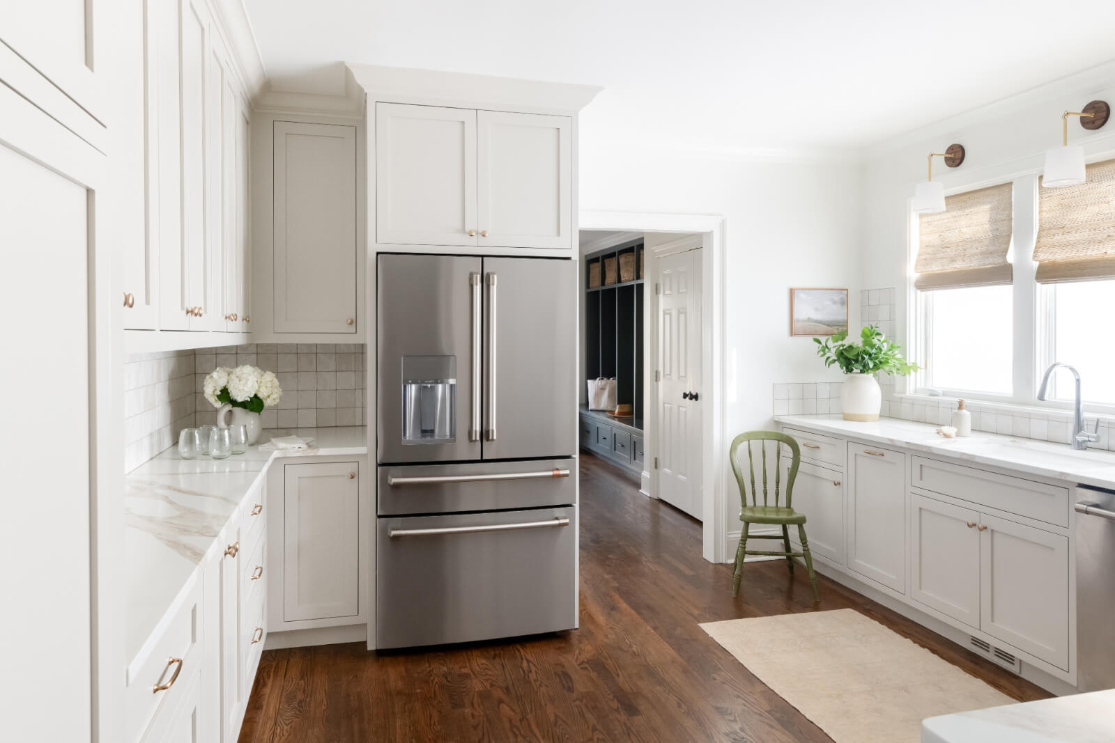 Modern kitchen designed by Elizabeth Burch of Home Interiors in Nashville, featuring a stainless steel refrigerator, white cabinetry, marble countertops, wooden flooring, and light streaming through a window with woven shades. A green chair and potted plant add vibrant accents.