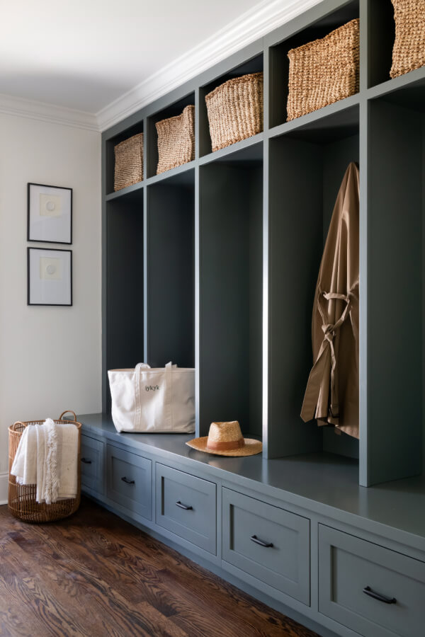 Entryway with dark green built-in cubbies, some containing wicker baskets. Designed by Elizabeth Burch Interiors, this Nashville home feature includes two framed pictures on the wall, a bag and hat on the bench, a hanging coat, and a basket with blankets on the floor.