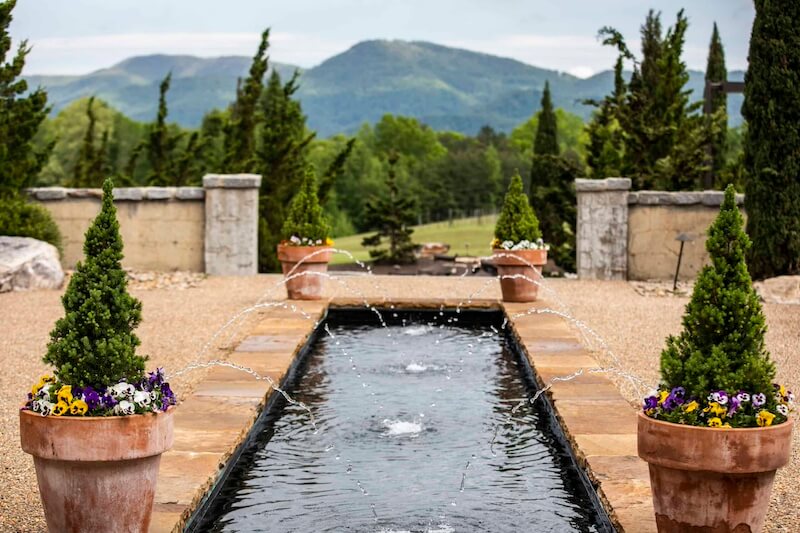 A long, rectangular water fountain with spouts is flanked by potted plants and flowers, set in a picturesque South Carolina garden with mountains visible in the background.