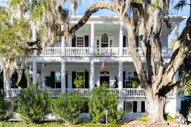 Large, white two-story house with a wraparound porch and columns, surrounded by greenery and a large tree with hanging moss. An American flag is displayed on the porch, evoking the charm of South Carolina living.