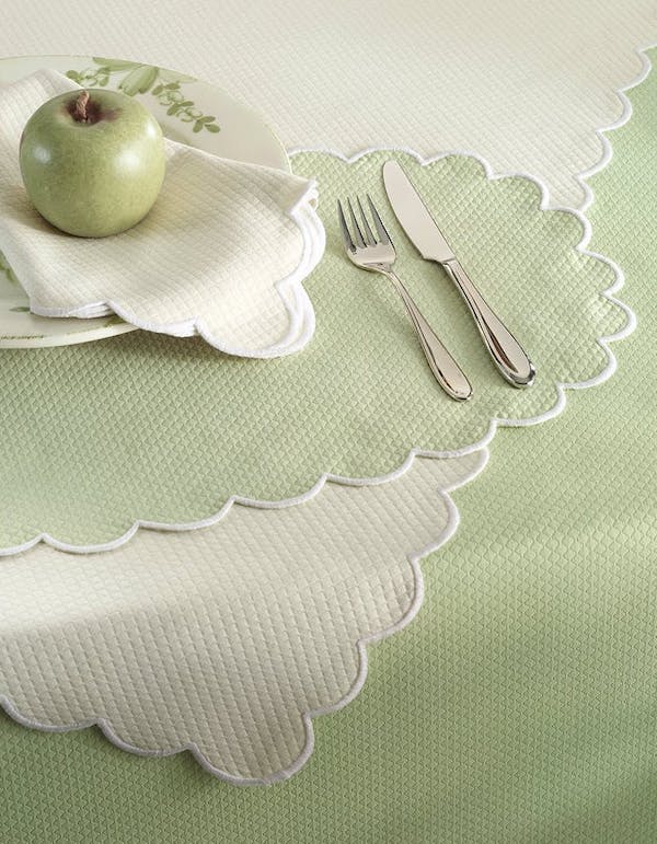 A plate with a green apple and a napkin is set on a scalloped-edge placemat, alongside a fork and knife, on a pale green textured tablecloth—perfect for those who appreciate simple luxe loves.