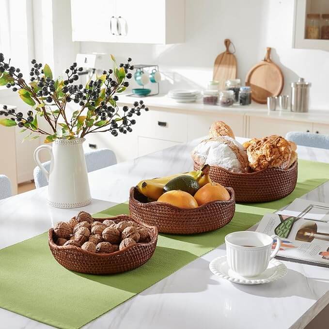 A piece of home decor, this kitchen table features a green runner, three rattan wicker baskets containing mixed fruit, bread, and nuts, a white vase with green and black berries, a coffee cup on a saucer, and an open book.