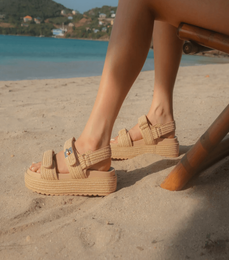 A person seated on a wooden chair on a sandy beach, wearing beige, woven platform sandals with the ocean and greenery visible in the background, creating a perfect blend of natural decor and Rattan furniture elements.