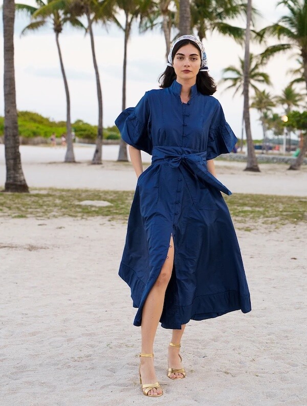 A woman wearing a blue belted dress and yellow sandals stands on a sandy beach with palm trees in the background, radiating luxe loves style.