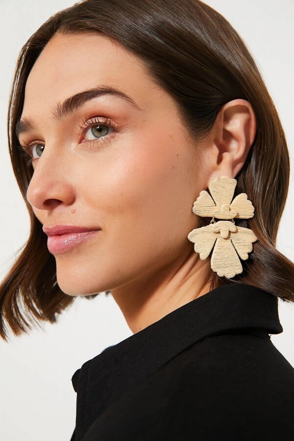 A woman with short brown hair is wearing a black top and large rattan flower-shaped earrings, facing left with a neutral expression.