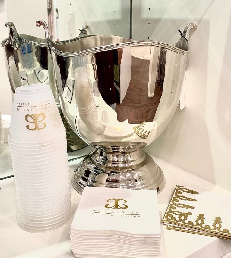 A large silver ice bucket is placed on a counter alongside a stack of white plastic cups and square coasters, all featuring the same gold monogram logo—perfect elements for expensive gift ideas.
