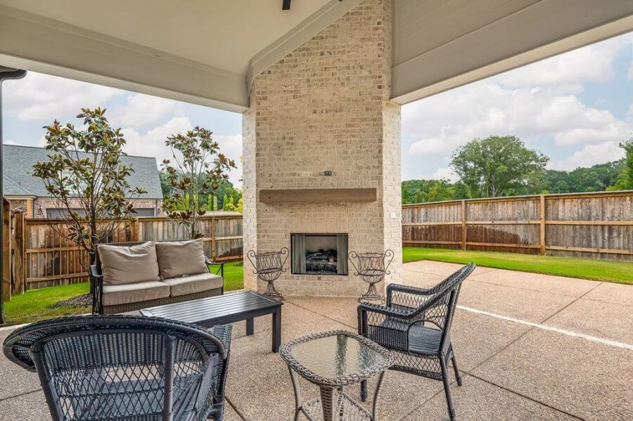 Relax on the covered patio at 140 Magnolia Ridge Drive in Rossville, Tennessee, featuring wicker furniture and a glass-topped table by a brick fireplace, all overlooking a fenced backyard with lush grass and trees.