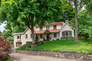 A two-story house at 79 Valley Road features a beige and white exterior, red roof, and a surrounding lawn with lush trees. The driveway leads to an attached garage on the left side, enhancing this Louisville home's charm.