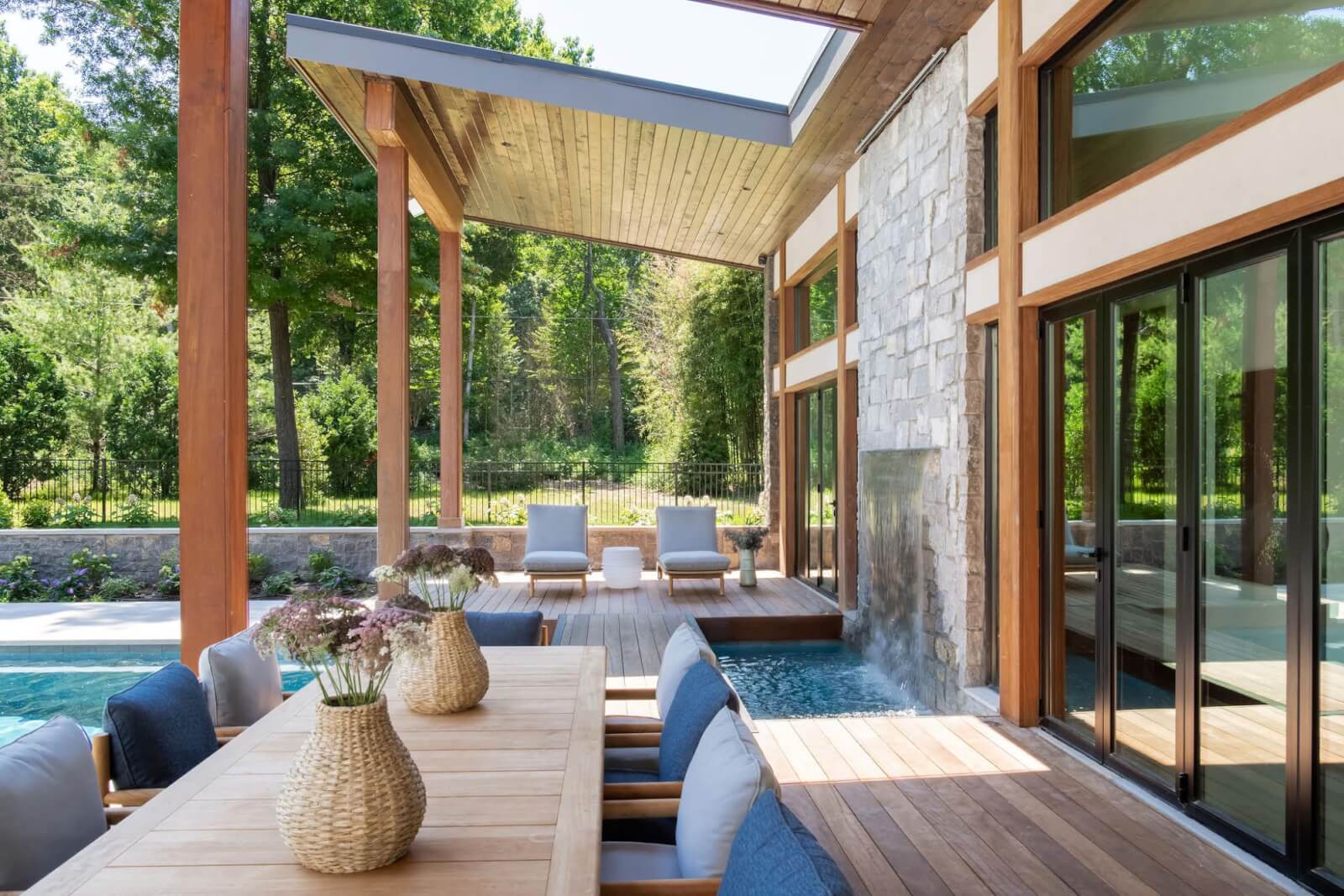 Modern patio with a wooden dining table and chairs, wicker vases, a stone waterfall, and two lounge chairs by the pool. Surrounded by greenery and large windows designed with insights from the best interior designers in Nashville.
