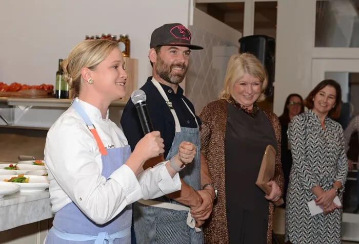 Three people, including Chef Lindsay Autry, are standing in a kitchen. The woman on the left is holding a microphone, the man is in the middle, and the woman on the right is holding a brown clutch. Several people are in the background.