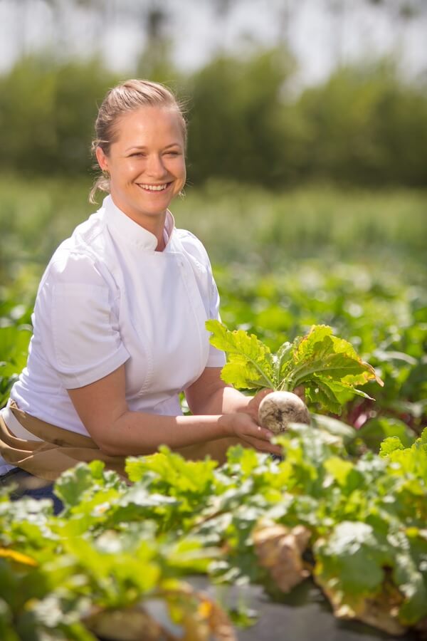 Chef Lindsay Autry, smiling in a white chef's uniform, stands in a lush green vegetable garden, holding a freshly picked beetroot.