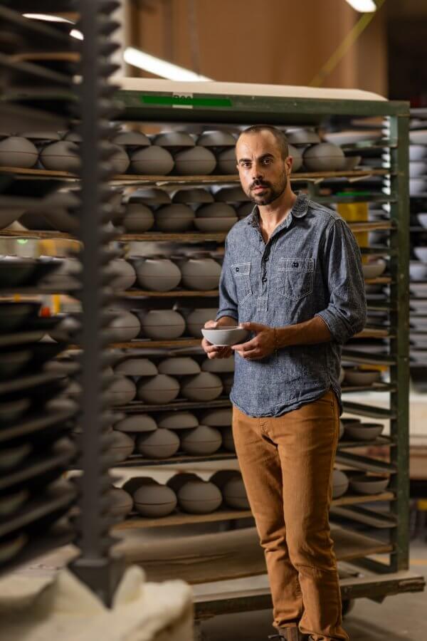 A man holding a ceramic bowl stands in front of shelves filled with East Fork Pottery in a workshop.