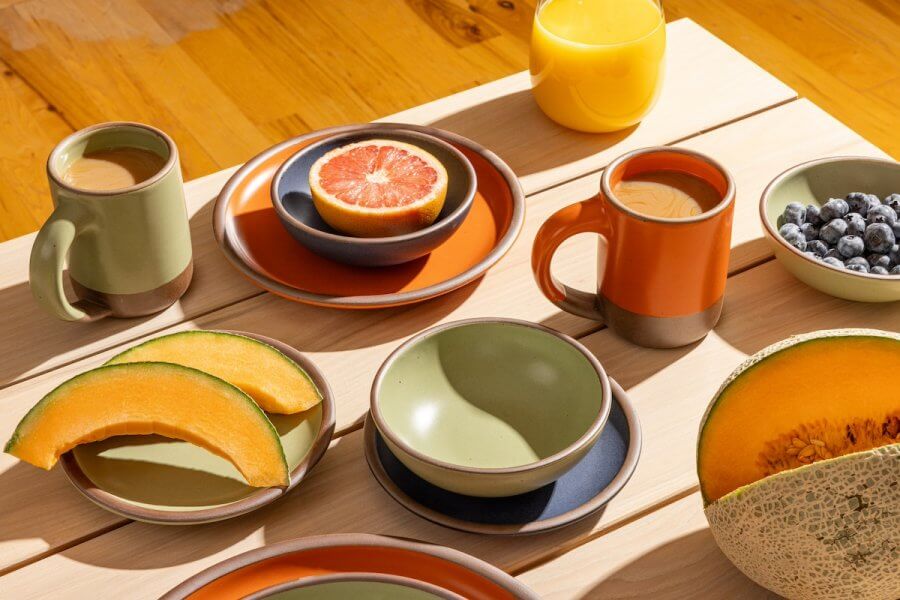 A wooden table with plates and mugs featuring a breakfast spread including melon slices, a grapefruit half, blueberries, and orange juice. The east fork pottery dishware is in earthy tones of green and orange.