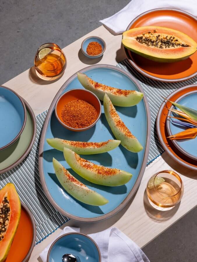 A table set with slices of melon sprinkled with chili powder on an East Fork pottery blue platter, halved papaya, small bowls of spice, glasses of drinks with lemon slices, colorful plates, and utensils.