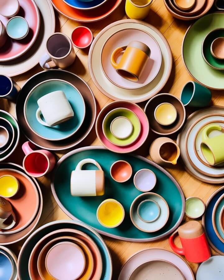 An assortment of colorful East Fork Pottery ceramic plates, bowls, and mugs arranged closely together, displaying a variety of sizes and shapes on a wooden surface.