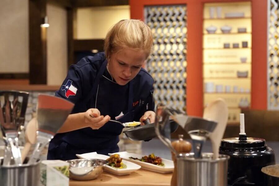 Chef Lindsay Autry, in a blue uniform, concentrates as she plates food in the kitchen. Various kitchen utensils and dishes are on the counter, while the background displays shelves with culinary equipment.