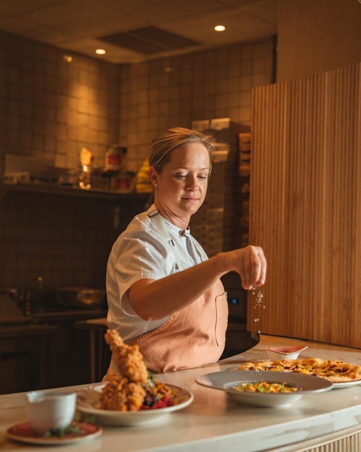 Chef Lindsay Autry is garnishing a dish in a kitchen, with several plated meals in front of her. She is wearing a white chef's jacket and a light pink apron. The kitchen setting includes tiled walls and shelves.