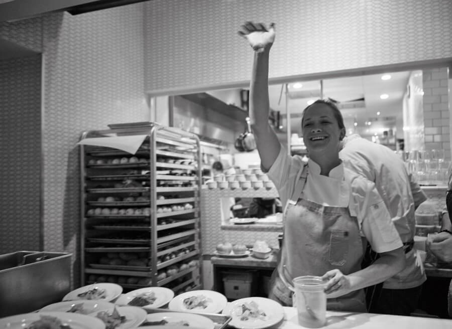 Chef Lindsay Autry waves with a smile in the bustling restaurant kitchen, with plates of food lined up on the counter and a rack of dishes in the background.