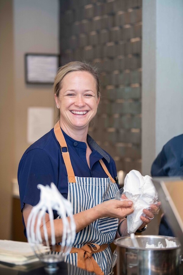 A person smiles while holding a bag of frosting. They are wearing a striped apron and standing in a kitchen setting. A mixing bowl and whisk are visible in the foreground.