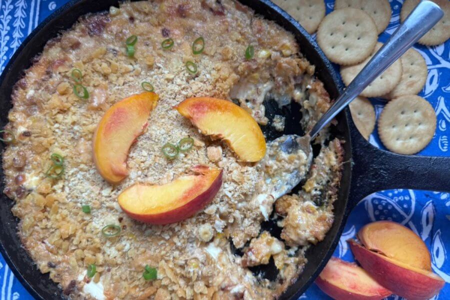 A skillet dish with a crumbly topping and garnished with peach slices, served alongside round crackers on a blue-patterned cloth, makes for the perfect Hot Cheesy Bacon Dip.