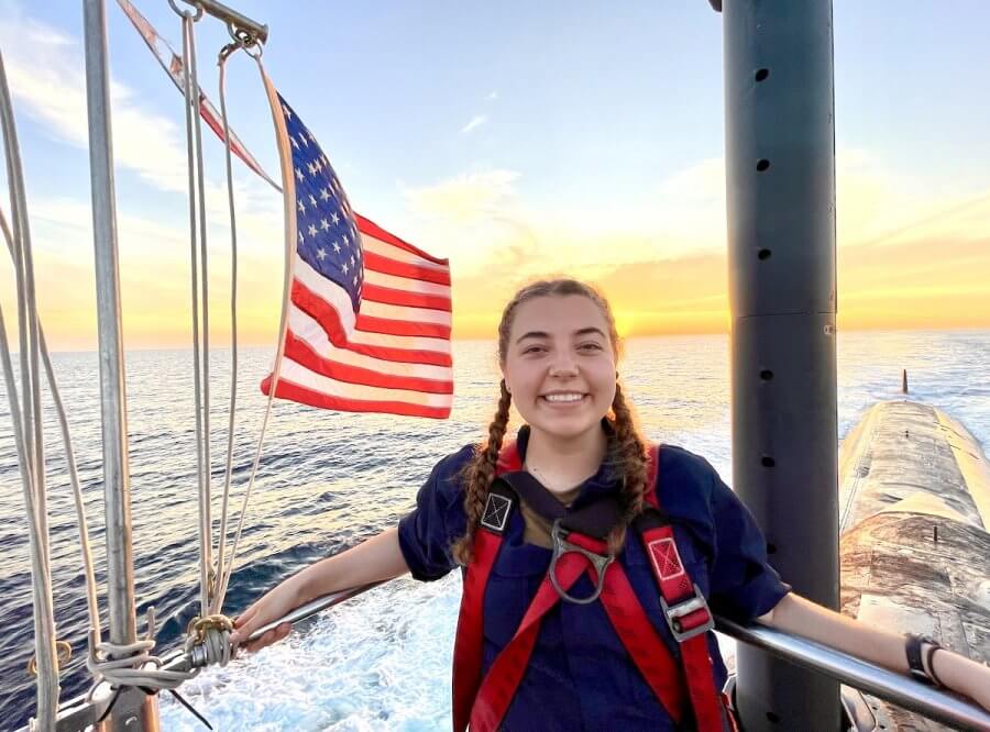 Reese on a submarine with an American flag in the background, under a sunset sky over the ocean. The scene reflects the pride and spirit of women at the naval academy.