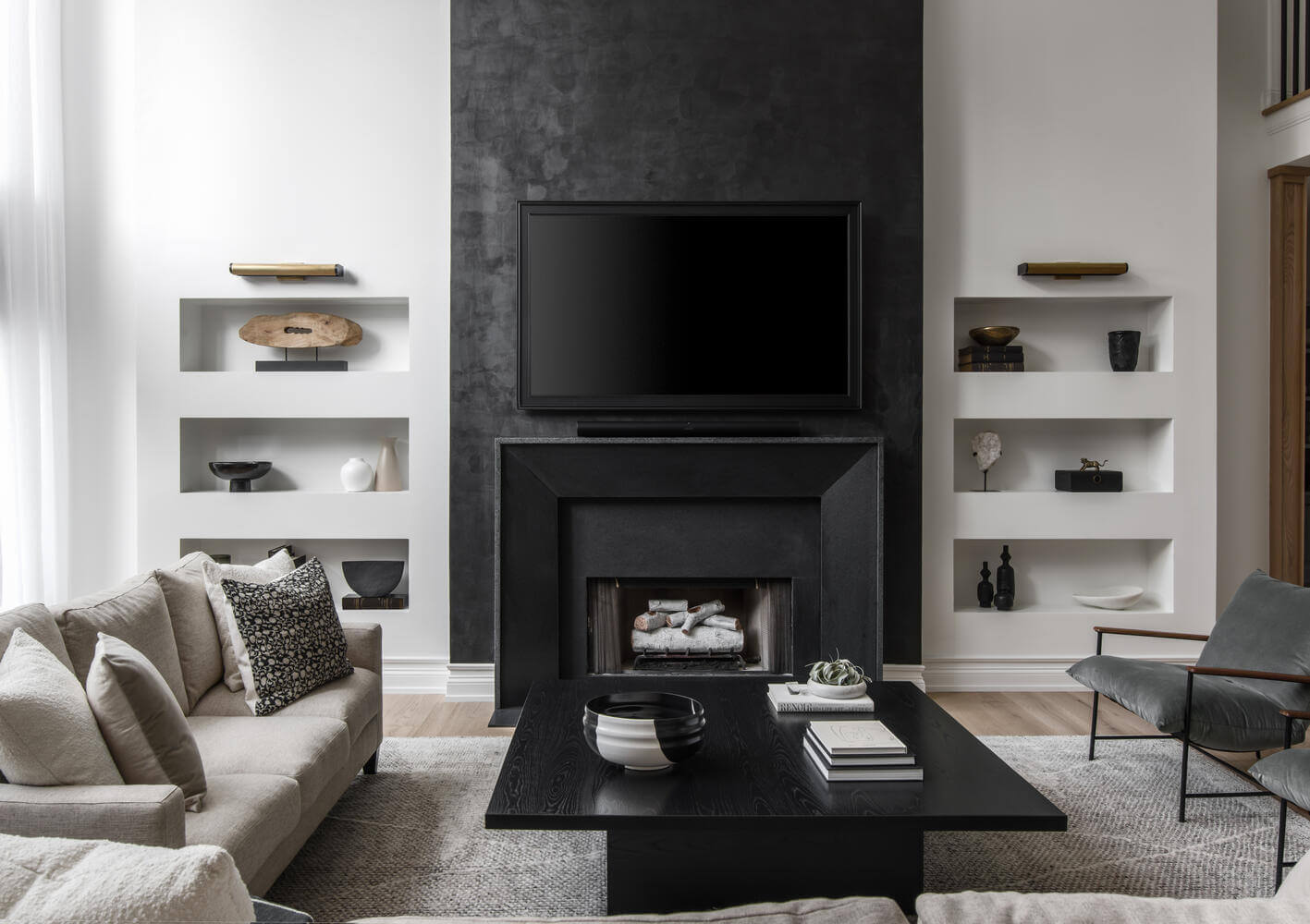 A modern living room designed by one of the best interior designers in Nashville features a black fireplace with a mounted TV, flanked by built-in white shelves. The room includes a gray sofa, black coffee table, and an armchair on a light-colored rug.