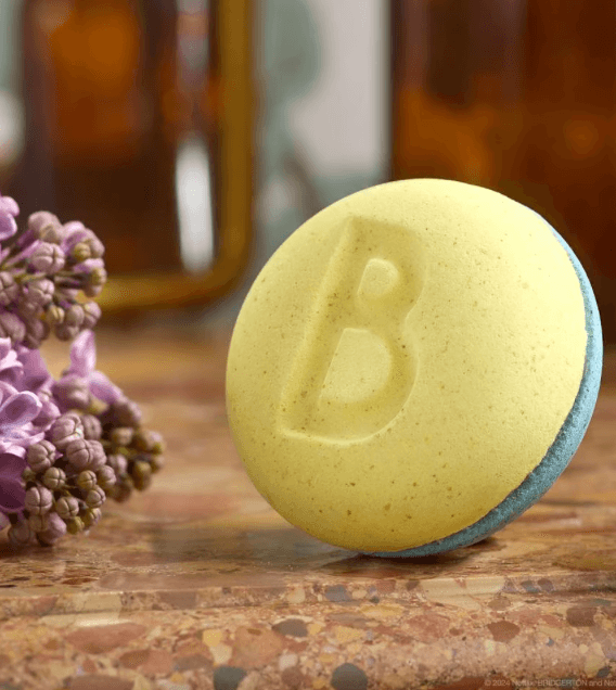 17 Birmingham FINDS Perfect for Summer - 12 A round yellow bath bomb with a "B" imprinted on it is placed on a marble surface next to purple flowers, perfect for adding a touch of summer luxury to your bathing routine.