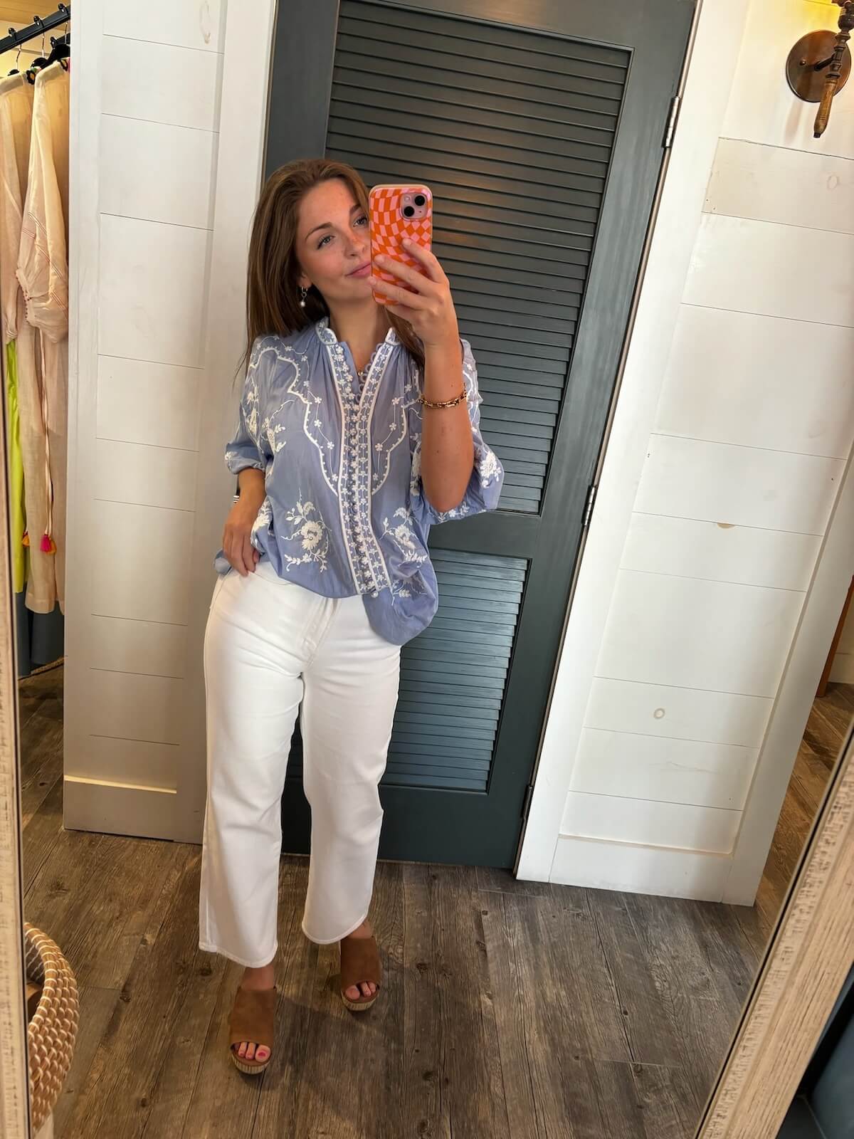 17 Birmingham FINDS Perfect for Summer - 3 A woman in a blue blouse and white pants takes a selfie in a fitting room, standing in front of a louvered door. She holds an orange patterned phone and wears brown heeled sandals.