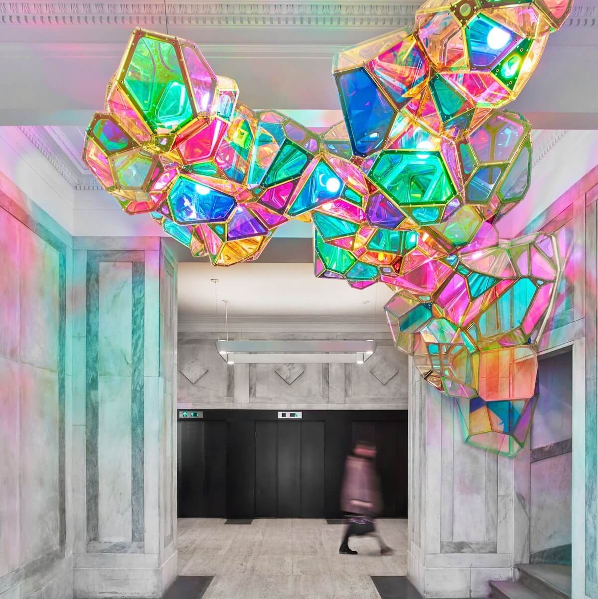 A colorful, geometric light installation hangs from the ceiling in a modern building lobby with marble walls and floors. A person walks by in the background.