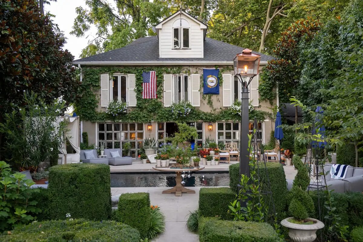 A lushly landscaped backyard with a rectangular pool, surrounded by greenery, patio furniture, and a two-story house adorned with ivy, American and state flags. 