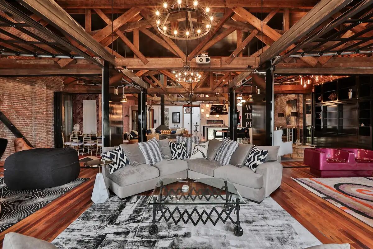 Spacious industrial-style living area with exposed wooden beams, large chandeliers, cozy sectional sofa, glass coffee table, and a modern kitchen in the background.
